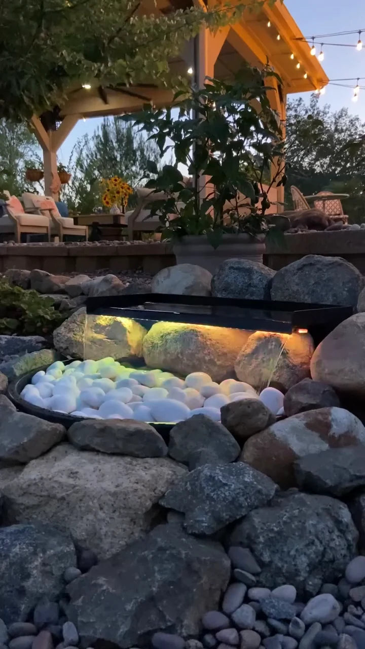 A glowing waterfall set into garden rocks and white pebbles, with a seating area and pergola behind it at dusk.